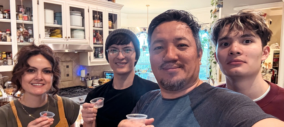Emma, Peter, Preston and Julian smiling in a cozy kitchen, each holding a small cup for a toast. The scene is warm and inviting, with cabinets and kitchenware visible.