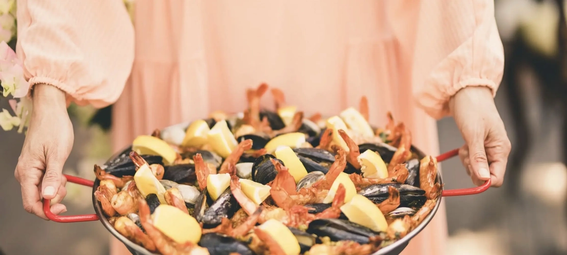 A person in a peach dress holds a large pan of vibrant seafood paella with shrimp, mussels, and lemon wedges, conveying a warm and inviting atmosphere.